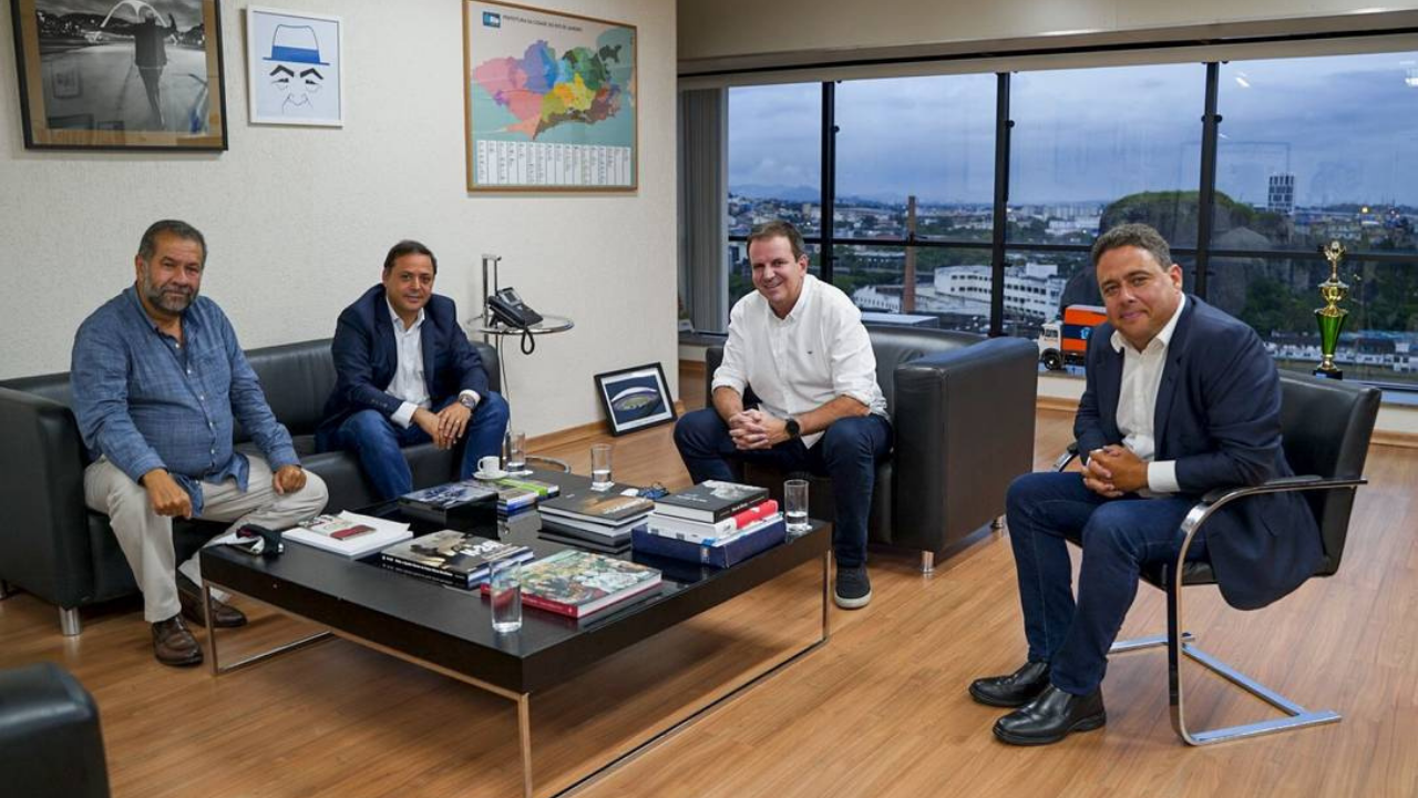 Foto de Eadurado Paes com Carlos Lupi e outros representantes na prefeitura do Rio. Há um centro com livros e revistas, um sofá, uma poltrona, piso de cerâmica. Na parede há uma fotografia não identificada e um mapa do Rio.