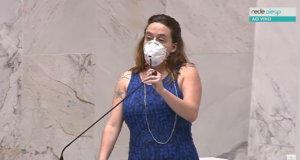 Foto de Isa Penna falando ao microfone na Alesp. Ela usa um vestido azul, máscara PFF2, cabelos lisos e soltes e um olhar firme.