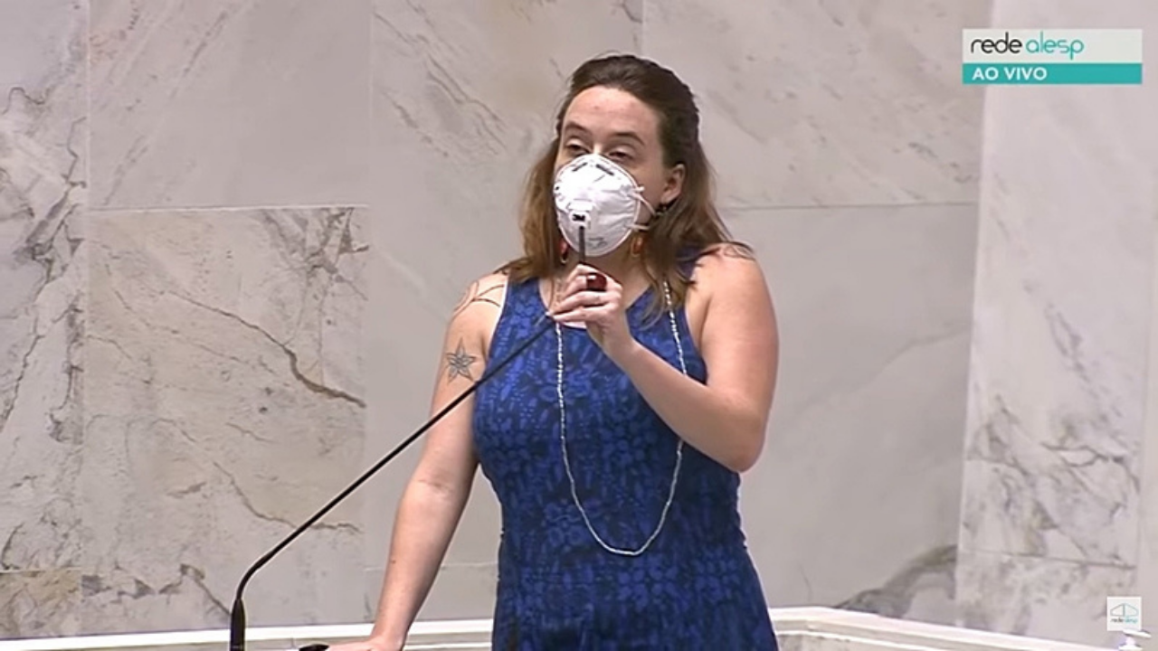 Foto de Isa Penna falando ao microfone na Alesp. Ela usa um vestido azul, máscara PFF2, cabelos lisos e soltes e um olhar firme.