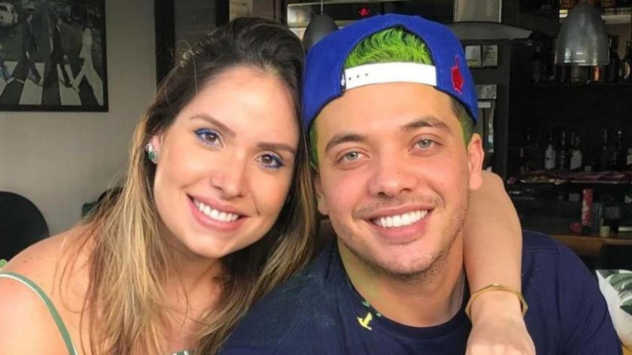 Foto de Wesley Safadão, sorrindo, com camisa azul e cabelo verde, ele usa um bone azul. Do lado esquerdo da foto está a companheira dele, Thyane Dantas, é loira, está sorrindo, ambos tem pele branca.