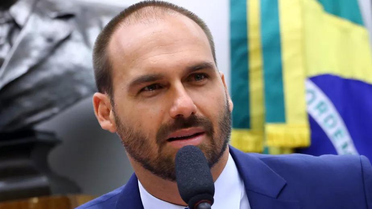 Eduardo Bolsonaro usa barba e fala ao microfone com olhar sério.