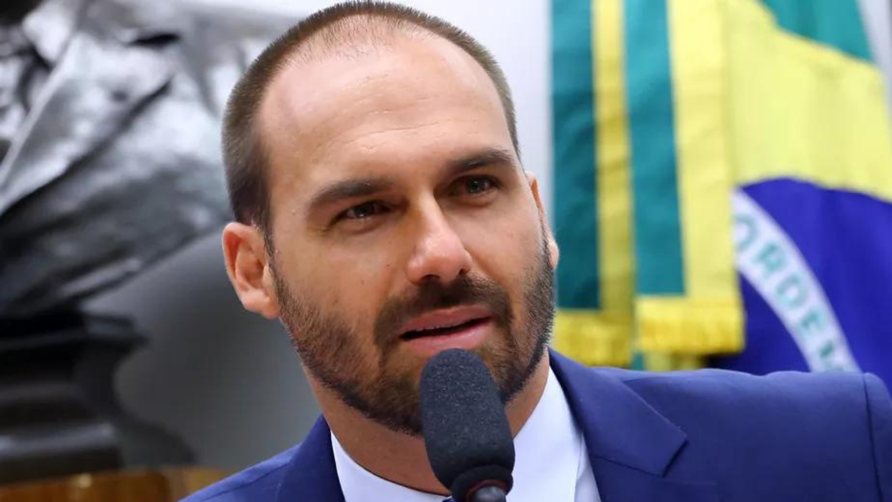 Eduardo Bolsonaro usa barba e fala ao microfone com olhar sério. 