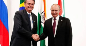 Bolsonaro em aproximação com Putin. Ele aperta a mão de Vladimir Putin, os dois usam ternos pretos, Bolsonaro com gravata azul e Putin com gravata vermelha. Ao fundo, há algumas bandeiras, incluindo a do Brasil.