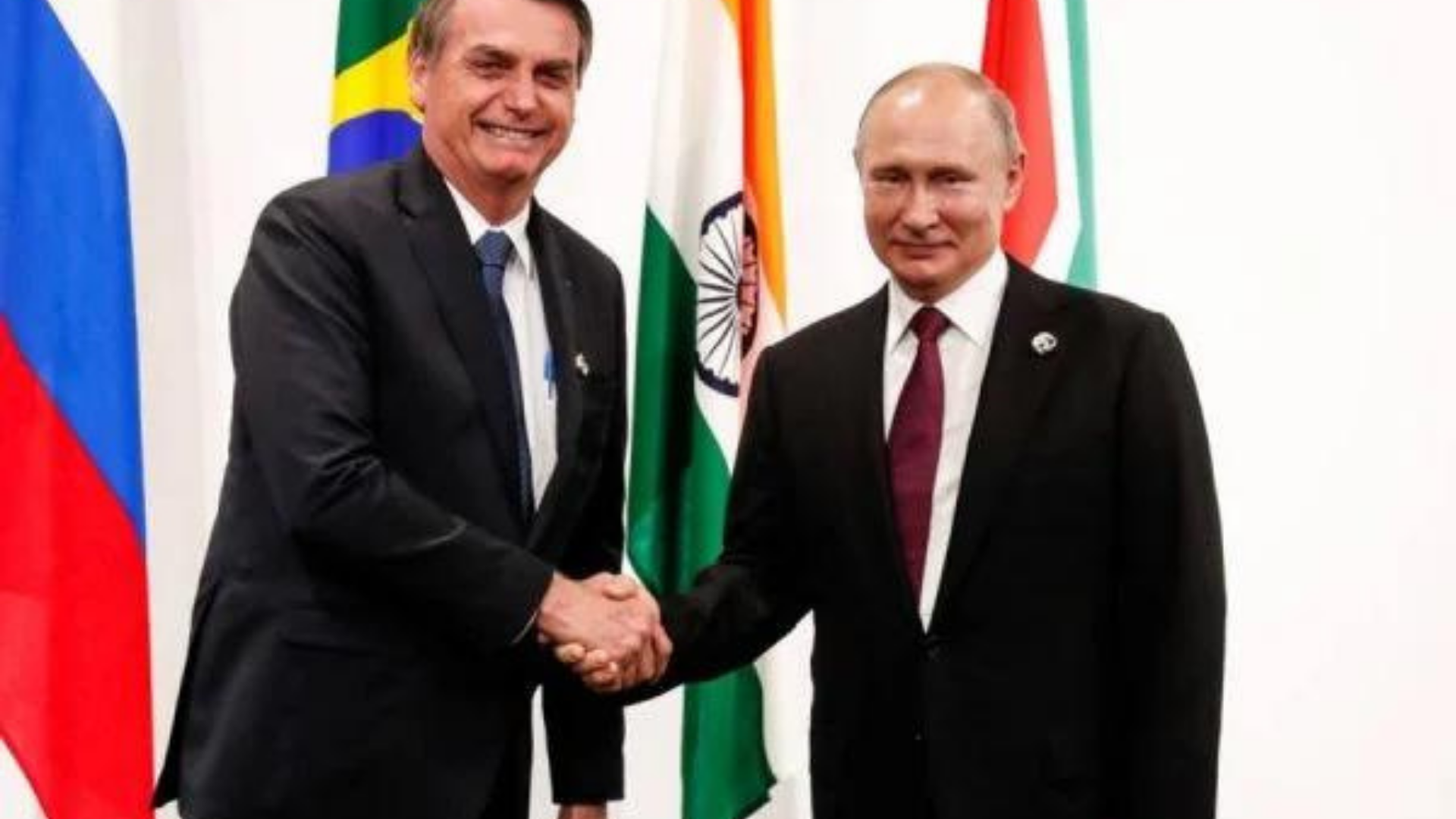 Bolsonaro irrita EUA em aproximação com rússia. Ele aperta a mão de Vladimir Putin, os dois usam ternos pretos, Bolsonaro com gravata azul e Putin com gravata vermelha. Ao fundo, há algumas bandeiras, incluindo a do Brasil.