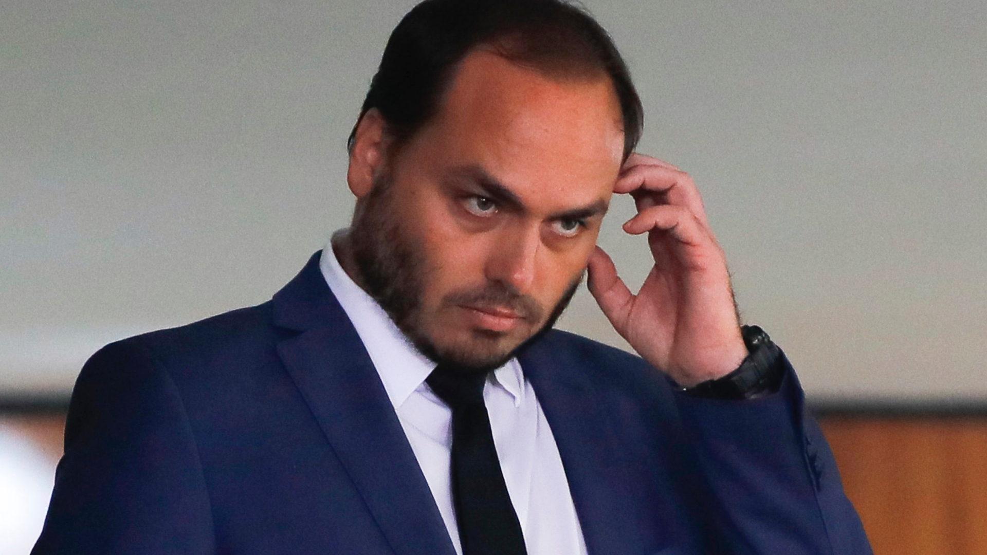 Carlos boslonaro usa cabelo e barba pretas, tem expressão cabisbaixa e olhar sério, com mão esquerda na altura da orelha