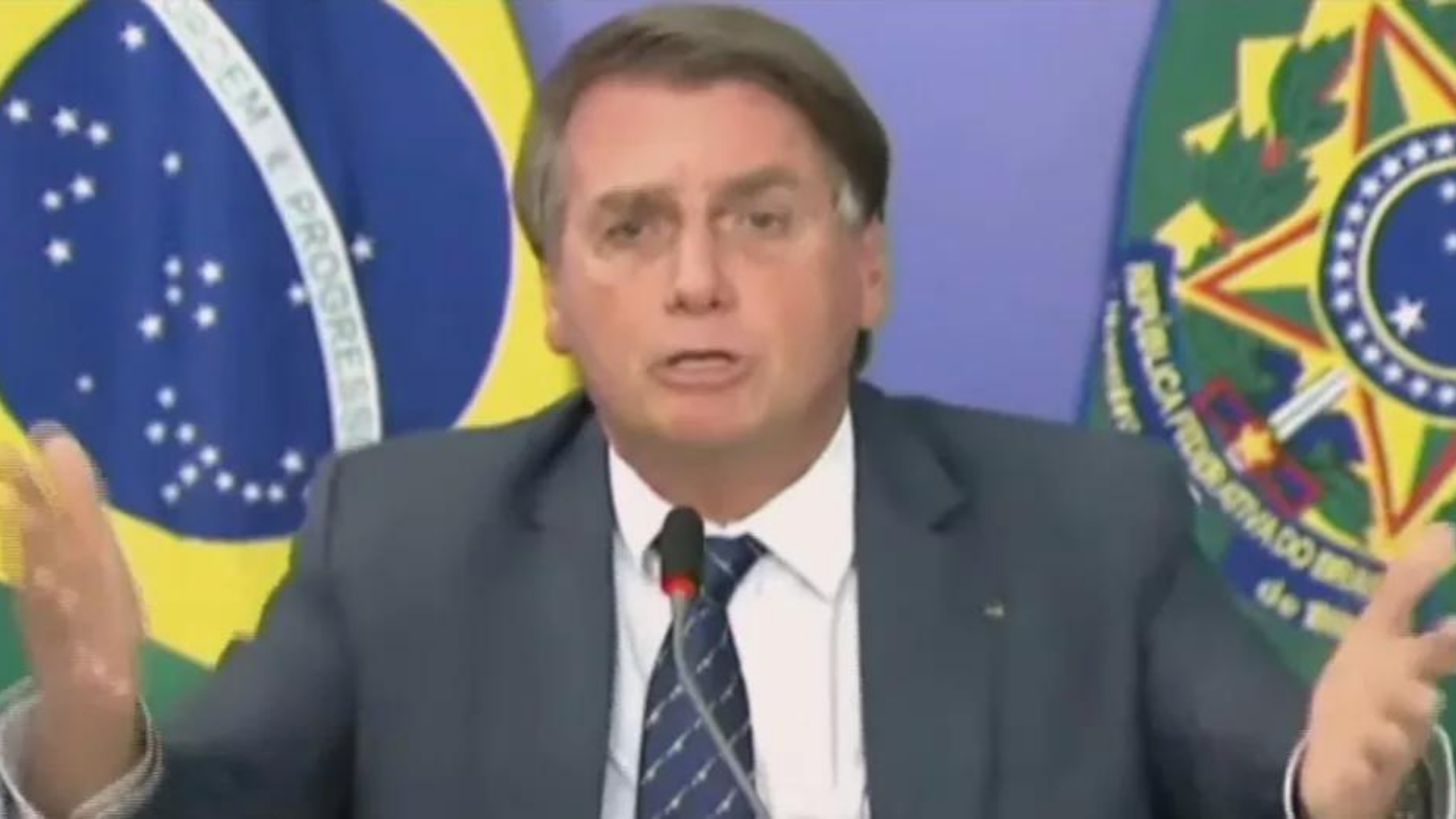 Bolsonaro surta e ataca esquerda. Foto do presidente usando terno cinza, fala ao microfone e tem os braços formando ângulo de 90°