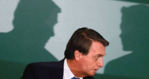 Bolsonaro diz a generais que já escolheu vice dos sonhos. Foto do Bolsonaro sentado pegando a lateral do rosto, ao fundo há a sombra de duas pessoas conversando.