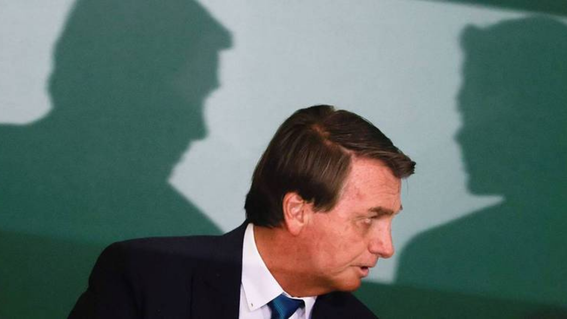 Bolsonaro diz a generais que já escolheu vice dos sonhos. Foto do Bolsonaro sentado pegando a lateral do rosto, ao fundo há a sombra de duas pessoas conversando.
