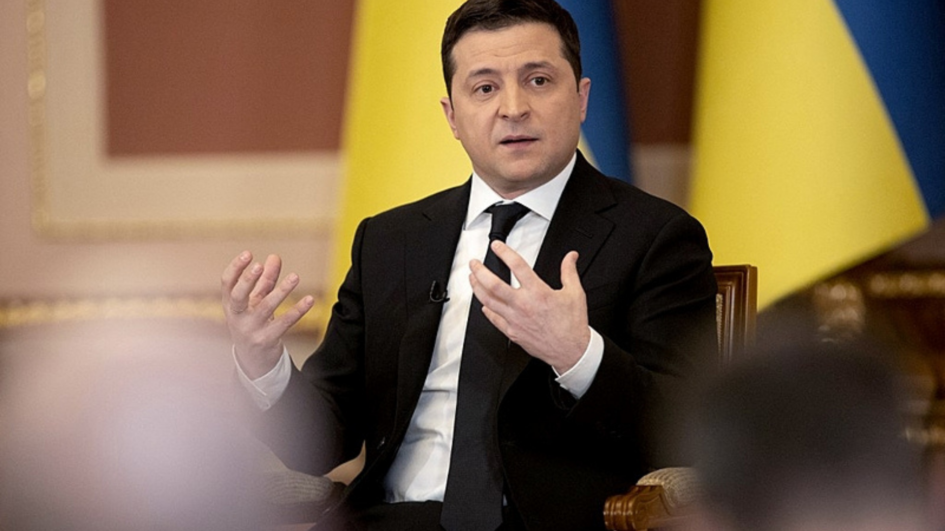 Zelensky diz que Rússia irá tomar o país na spróxima shoras. Ele aparece em foto com terno preto, expressão séria e gesticula com as mãos.