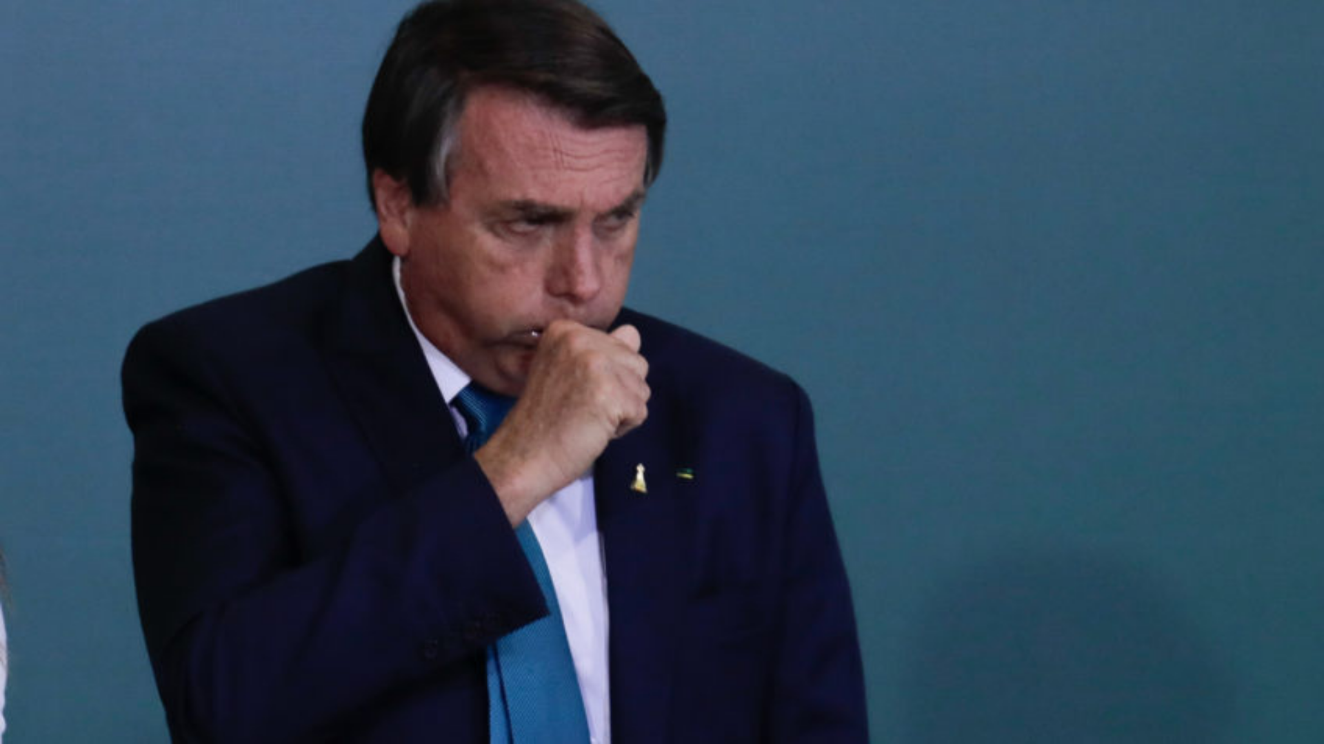 Bolsonaro mente sobre conversa com Putin. Bolsonaro usa terno e está tossindo com a mão na boca.