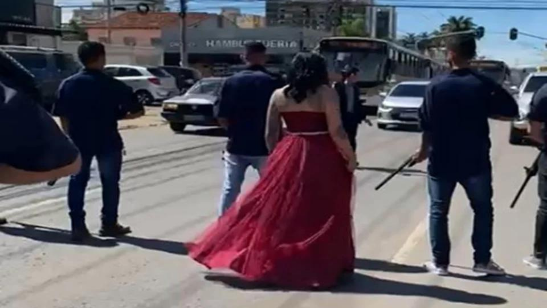 Homens com fuzis param trânsito para dama de vermelho passar. Mulher usando vestido vemelho para no meio da rua junto com homens armados com fuzis.