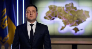 Presidente da Ucrânia faz apelo aos russos. Ele usa terno preto, tem expressão séria no rosto e usa a bandeira e um mapa da Ucrânia ao fundo.