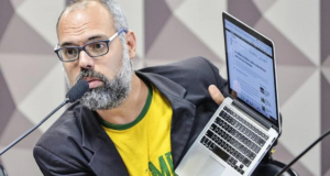 Allan dos Santos ataca Moraes. Blogueiro usa camisa verde e amarela com terno preto, óculos, barba e cabelo grisalhos. Segura laptop com as mãos