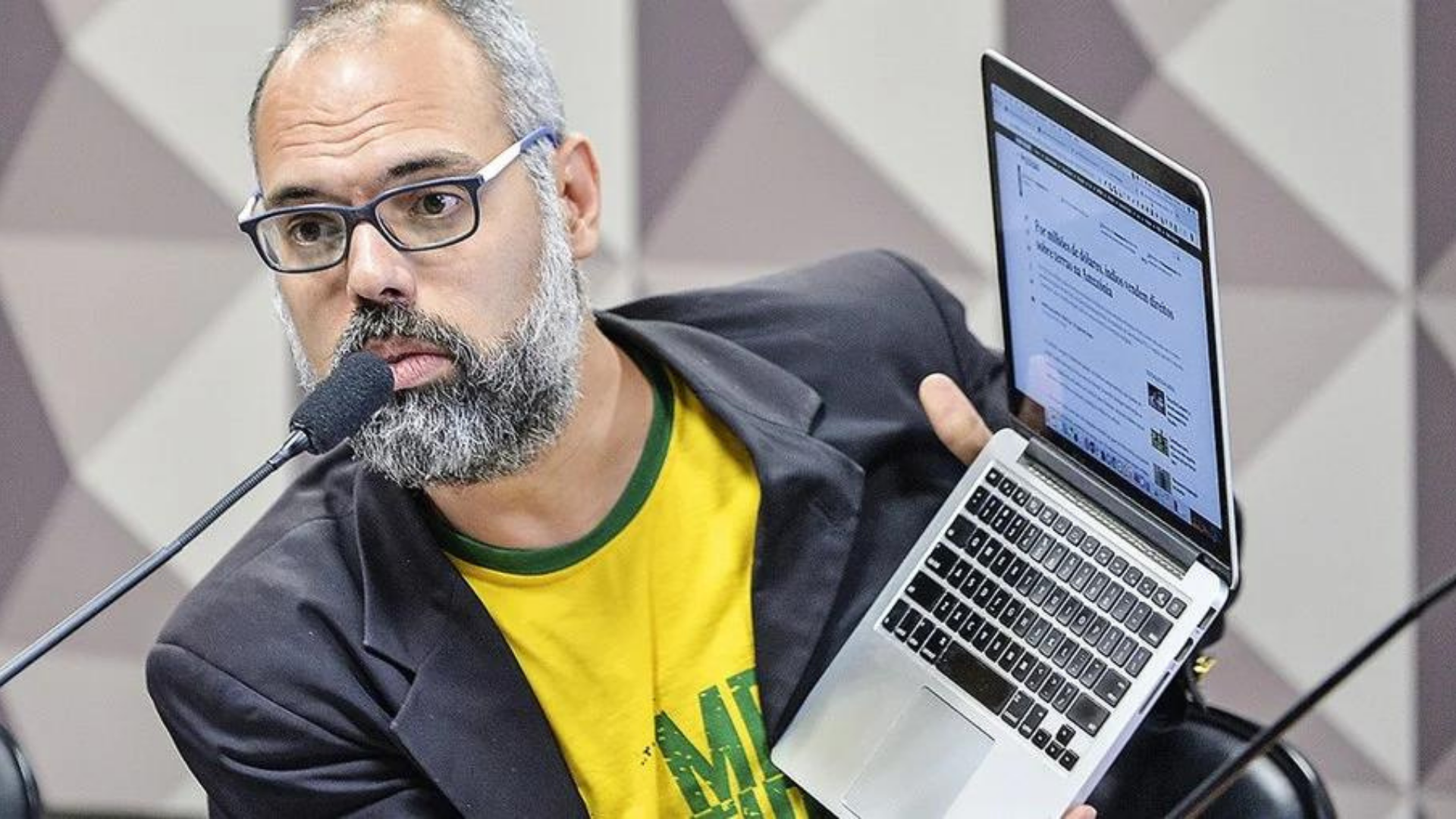 Allan dos Santos ataca Moraes. Blogueiro usa camisa verde e amarela com terno preto, óculos, barba e cabelo grisalhos. Segura laptop com as mãos