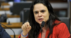 Janaína Paschoal sobre Bolsonaro não apoia-la ao Senado: "Tem medo de mim". Ela tem expressão facial de raiva, está apontando o dedo indicador direito, tem os cabelos soltos e usa um vestido laranja.