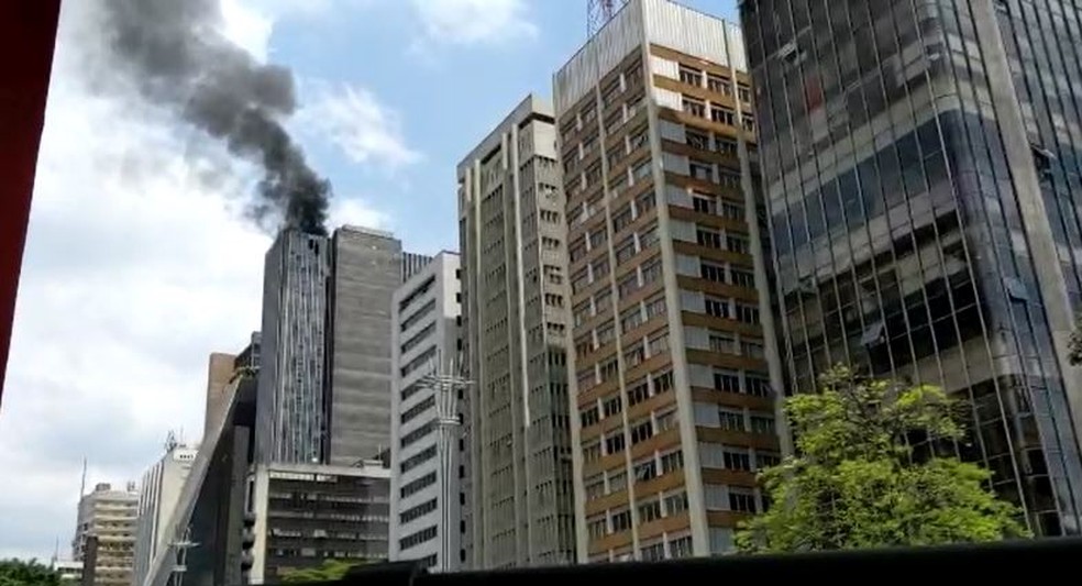 Imagem um pouco mais distante, onde é possível ver outros edifícios ao lado, mostra fumaça preta saindo do prédio que pegou fogo.