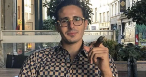 Shimon Hayut sentado, olhando para frente, com um charuto na mão. Ele é branco, usa óculos e uma camisa xadrez.