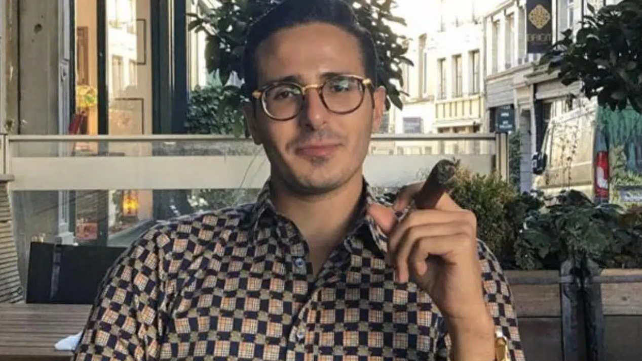 Golpista do Tinder, Shimon Hayut sentado, olhando para frente, com um charuto na mão. Ele é branco, usa óculos e uma camisa xadrez.