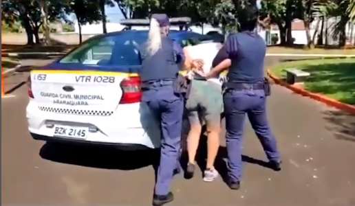 Mulher negacionista sendo conduzida à viatura após tentar resistir à prisão, em Araraquara. Ela mantém as mãos nas costas, enquanto é levada por dois policiais.