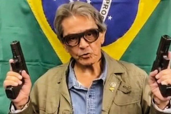 Roberto Jefferson com bandeira do Brasil pendurada atrás dele e segurando uma arma em cada mão.