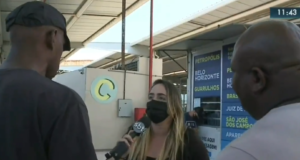 A repórter está no centro da imagem, com o microfone na mão, enquanto dois homens ficam entre ela e a câmera, tentando impedir o trabalho da reportagem.