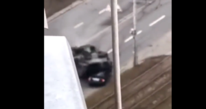 Momento em que um tanque de guerra atropela um carro, em uma estrada. A imagem é aérea, feita de um prédio.