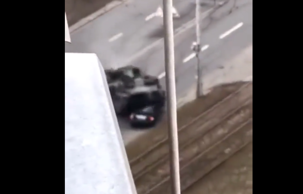 Momento em que um tanque de guerra atropela um carro, em uma estrada. A imagem é aérea, feita de um prédio.