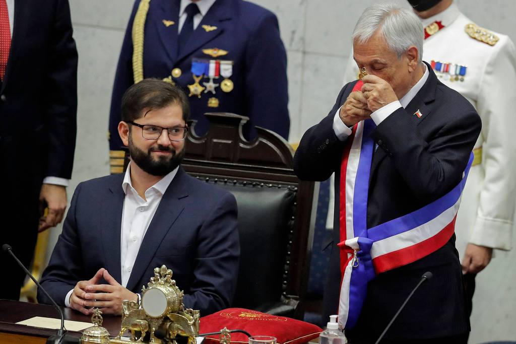 Gabriel Boric sentado e sorrindo. Ao seu lado, Sebatián Piñera ainda com faixa presidencial beija um escudo que é o símbolo do poder no Chile.