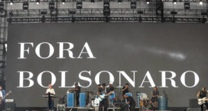 Mensagem "Fora Bolsonaro" em projeção no palco do festival musical Lollapalooza, enquanto uma banda se apresenta.