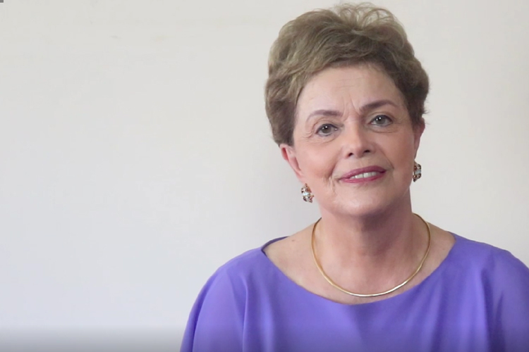 Dilma posando para foto, com camisa roxa e fundo branco. Ela sorri.