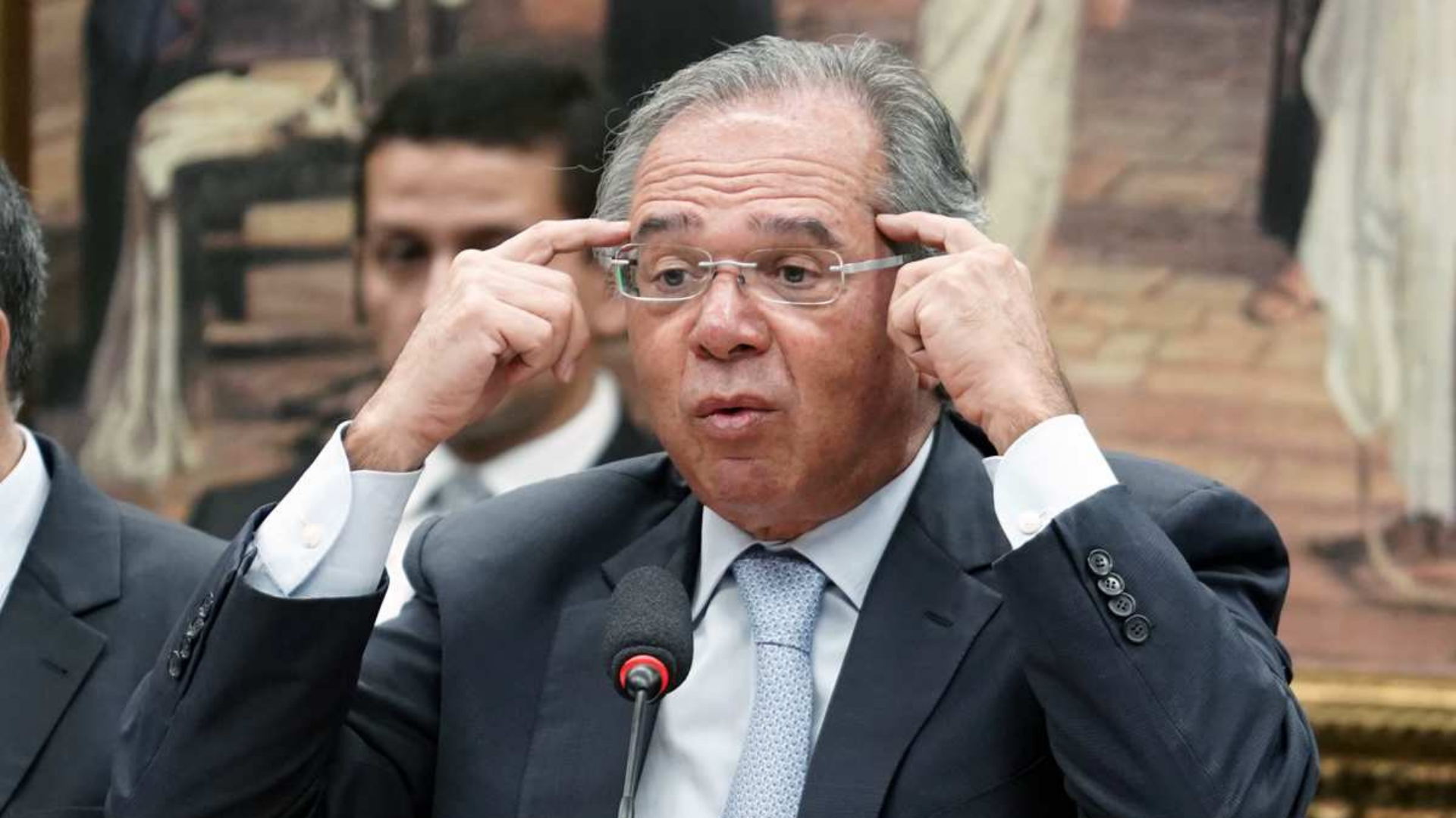 Paulo Guedes apontando para a própria cabeça, como se estivesse pensando. Ele está sentado, de óculos, terno e gravata e com um microfone à sua frente.