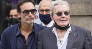 Alain Delon pede ao filho que lhe lhe ministre eutanásia. Foto de Alain Delon e do filho, ambos com pele branca, usando óculos de sol e ternos na cor preta e azul marinho, respectivamente.