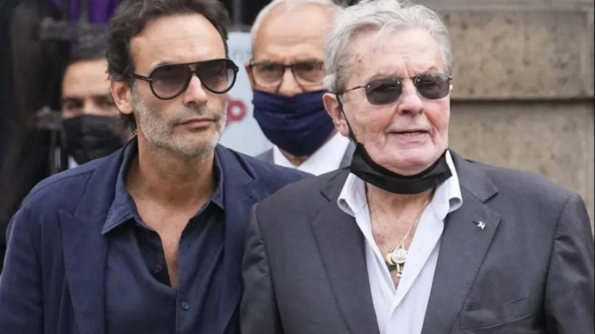 Alain Delon pede ao filho que lhe lhe ministre eutanásia. Foto de Alain Delon e do filho, ambos com pele branca, usando óculos de sol e ternos na cor preta e azul marinho, respectivamente.