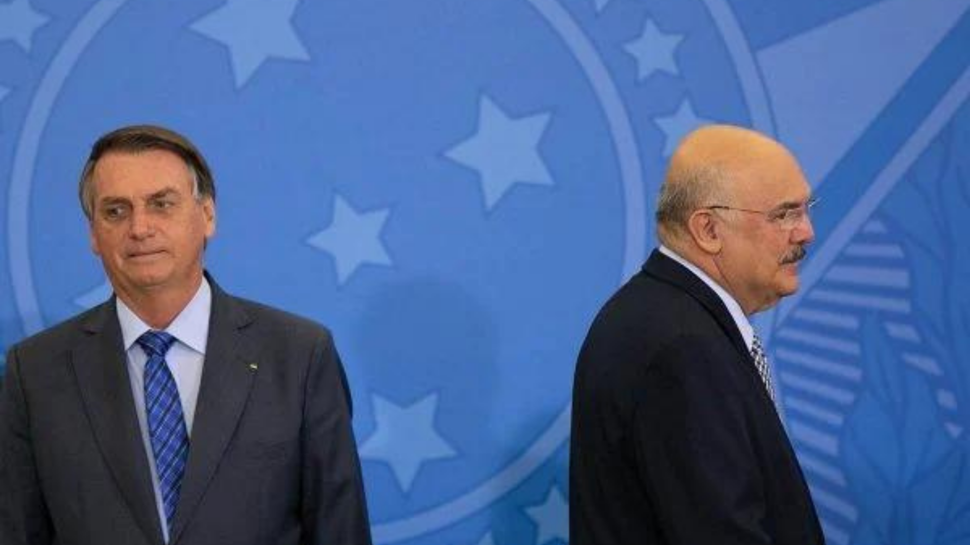 Internação de Bolsonaro ajuda a ofuscar crise no governo. Foto de Bolsonaro com olhar desconcertado e o ministro Milton Ribeiro de costas para ele.