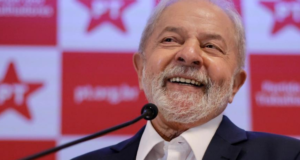 Lula sorrindo, falando ao microfone. Logo do PT ao fundo; ele lidera pesquisa Datafolha