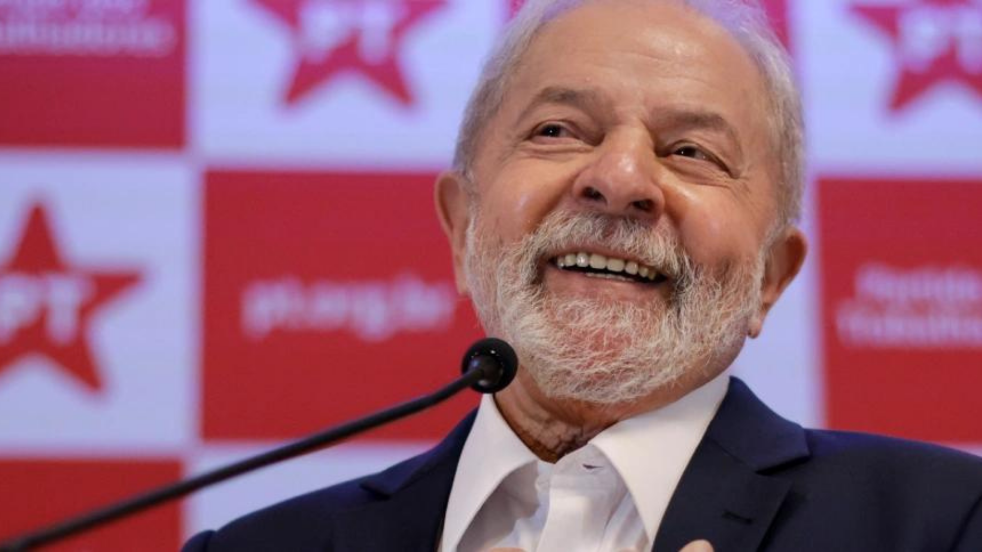 Lula sorrindo, falando ao microfone. Logo do PT ao fundo; ele lidera pesquisa Datafolha