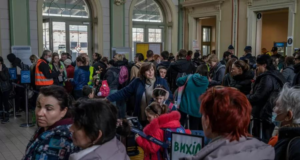 Novos refugiados ucranianos chegam ao Brasil. Foto dos imigrantes em um salão utilizando casacos e expressões sérias.
