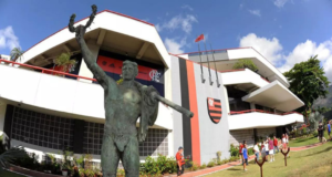 Homem tenta estuprar criança de 10 anos em vestiário do Flamengo. Foto da entrada do Clube, um prédio com arquitetura contemporânea, símbolo do Flamengo e uma estátua ao lado de fora.