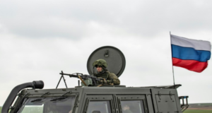 Putin autoriza convocação de soldados sírios para lutar na Ucrânia. Foto de um soldado dentro d eum tanque de guerra e uma bandeira da Rússia trmulando.