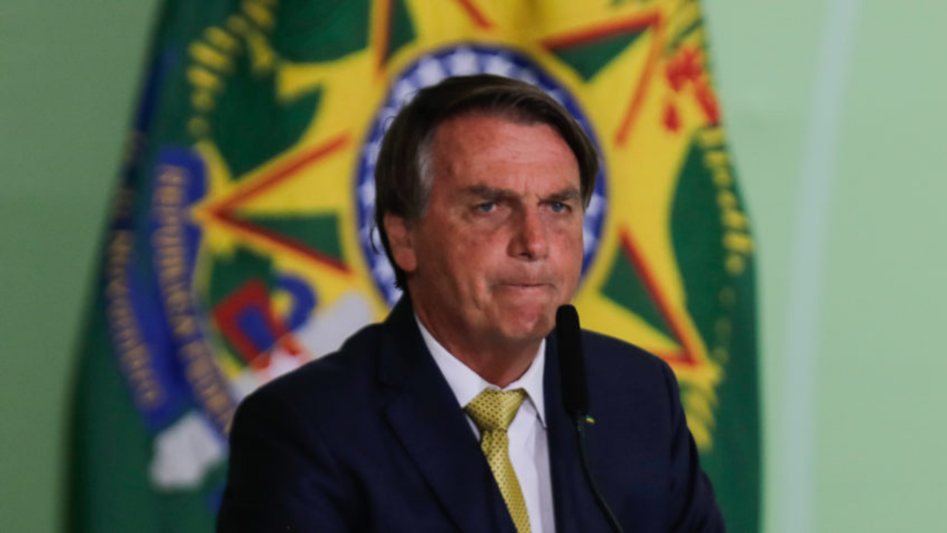 Bolsonaro conta a piada do dia: "Não tomamos decisões pensando nas eleições". Foto de Bolsonaro com expressão de preocupação falando ao microfone. 