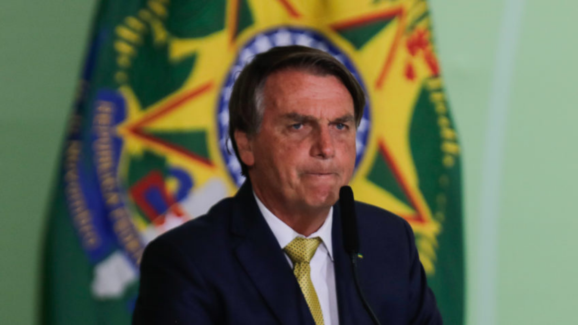 Bolsonaro conta a piada do dia: "Não tomamos decisões pensando nas eleições". Foto de Bolsonaro com expressão de preocupação falando ao microfone.