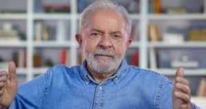 Lula teme assassinato por parte de bolsonaristas. Ele tem os braços levantados na altura do ombro, olhar sério e veste uma camisa jeans.