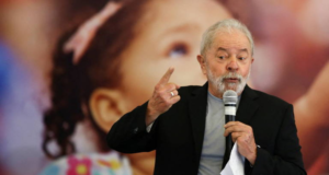 AO VIVO: Lula discursa em evento no México. Foto do ex-presidente falando ao microfone, usando terno preto e camisa listrada cinza com branco. Cabelos e barba brancos.