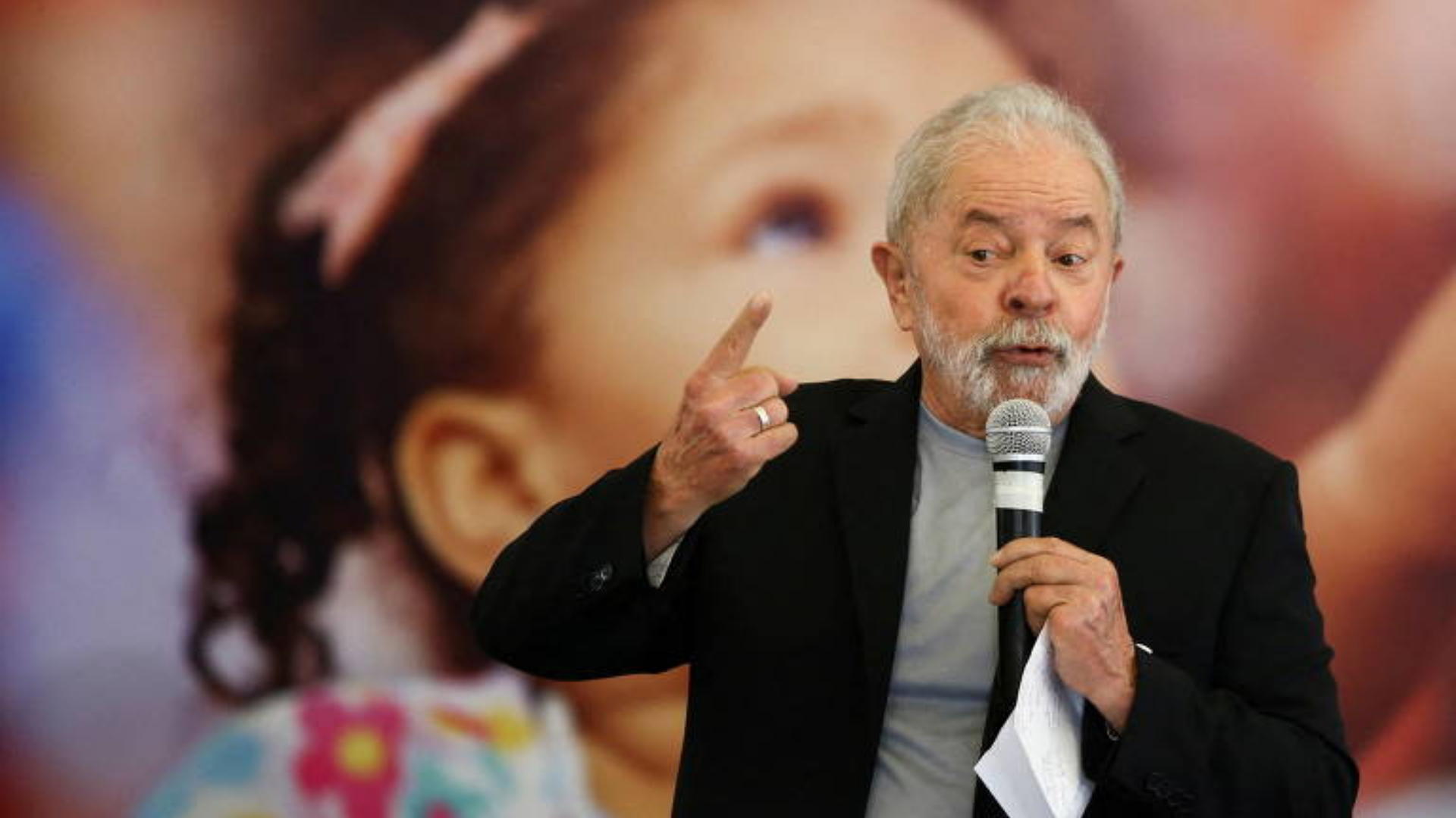 AO VIVO: Lula discursa em evento no México. Foto do ex-presidente falando ao microfone, usando terno preto e camisa listrada cinza com branco. Cabelos e barba brancos.