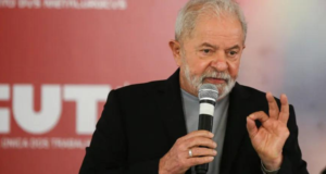 Caiu na rede: PowerPoint mostra que Lula será indenizado. Foto de Lula fazendo sinal de 'foda" com a mão esquerda, enquanto segura um microfone com a outra mão.