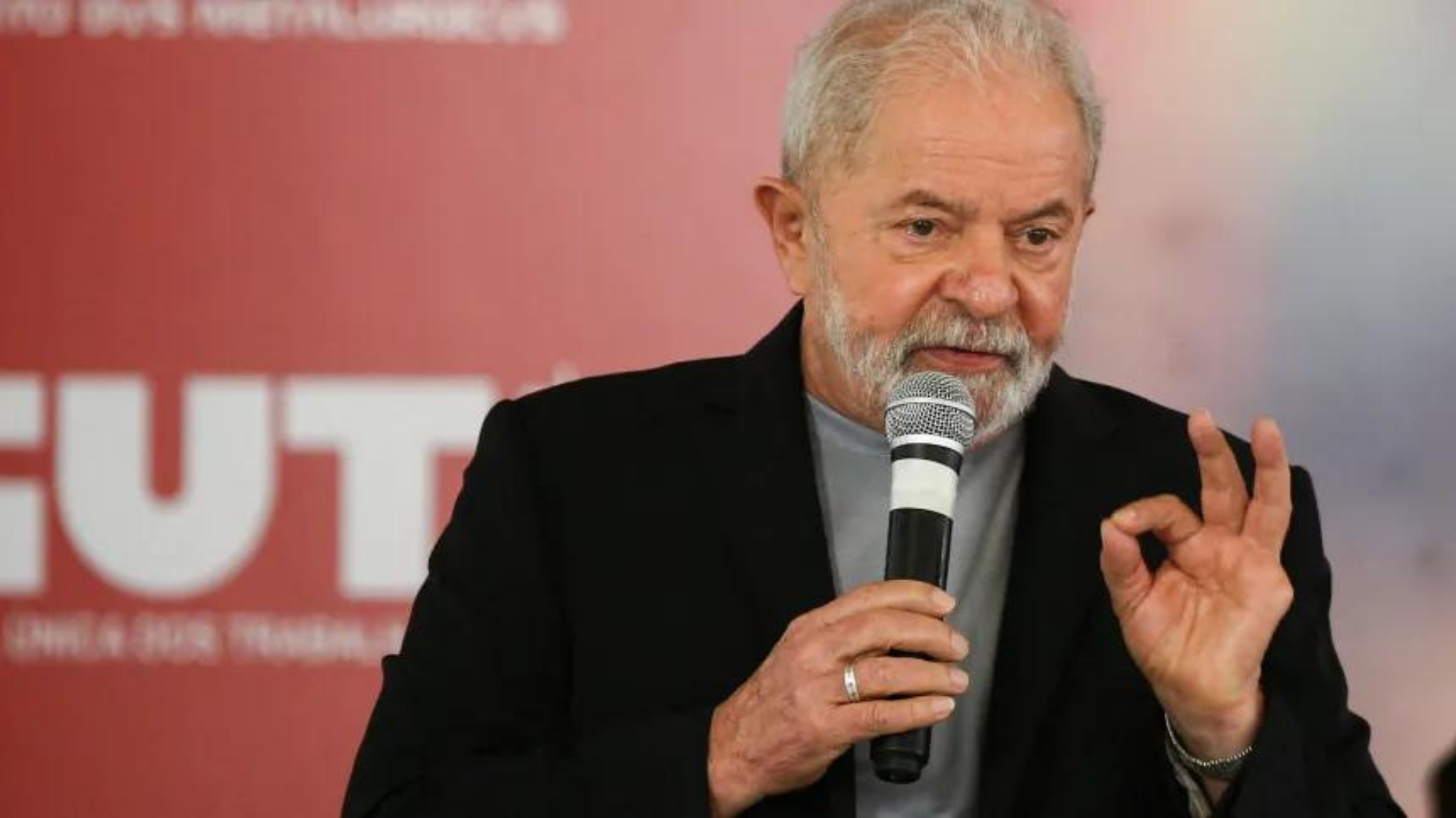 Caiu na rede: PowerPoint mostra que Lula será indenizado. Foto de Lula fazendo sinal de 'foda" com a mão esquerda, enquanto segura um microfone com a outra mão.
