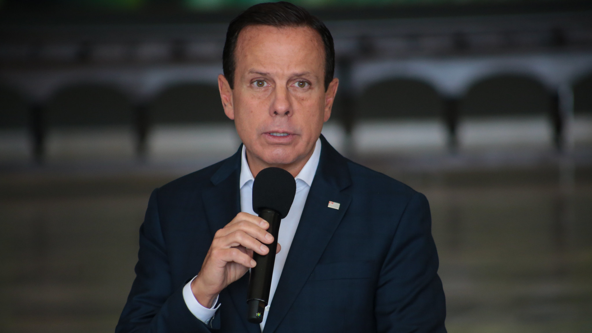 Nanico nas pesquisas, Doria chama de "golpista" quem quer tirá-lo da disputa. João Doria tem expressão de desânimo enquanto fala ao microfone. Ele está vestindo um terno azul marinho.