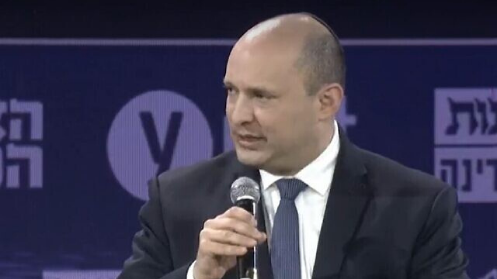 Premiê de Israel critica Zelensky por comparação com o Holocausto. Foto de Benneti usando terno azul e falando ao microfone.