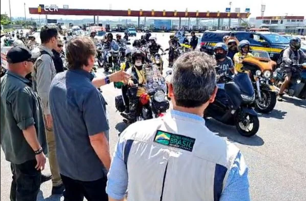 Bolsonaro e o ministro Tarcísio de Freitas, de costas, cumprimentando a motociata que passa em sua frente.