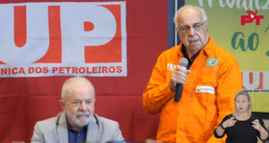 O ex-presidente Lula (PT) participando de debate com petroleiros. Ele está sentado, atrás dele há uma bandeira da FUP e ao seu lado um homem está em pé e fala ao microfone.